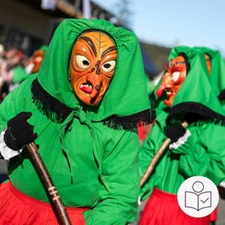 Verkleidete Hexen mit grünem Umhang. Die Hexe hat eine Holz·maske. Die Hexe hat einen Besen in der Hand. Rechts das SWR Logo für Leichte Sprache.