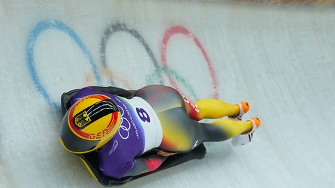 Ein Skeleton-Sportler rast durch die Eis·bahn. Er liegt auf dem Bauch auf seinem Schlitten. Er fährt mit dem Kopf voraus. 