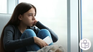 Ein Mädchen sitzt am Fenster. Sie hält ihr Handy in der Hand. Sie schaut traurig aus dem Fenster. Mehr Kinder und Jugendliche leiden wieder unter psychischer Belastung. Rechts das SWR Logo für Leichte Sprache.