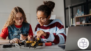 Das ist ein Beispiel·bild: Zwei Mädchen bauen einen Roboter auf einem Tisch zusammen. Rechts das SWR Logo für Leichte Sprache.