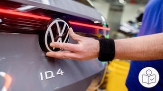 Ein VW-Mitarbeiter montiert das Firmenlogo auf der Rückseite eines Elektroautos. Rechts das SWR Logo für Leichte Sprache.