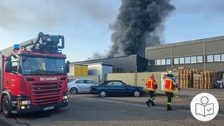 Ein Feuerwehrauto steht vor einer Industriehalle. Zwei Feuerwehrmänner laufen auf die Halle zu. Über der Halle steigt schwarzer Rauch auf. Rechts unten das SWR-Logo für Leichte Sprache.