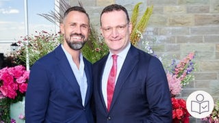Daniel Funke steht links. Jens Spehn steht rechts. Beide stehen vor einem Beet mit Blumen. Sie haben je einen Arm um den anderen gelegt und lächeln. Sie haben ein Kind von einer Leihmutter bekommen. Rechts unten das SWR-Logo für Leichte Sprache.