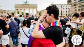 Auf einem großen Platz stehen zwei Männer und umarmen sich. Einer der beiden trägt ein Halstuch in Regenbogenfarben. Er wischt sich Tränen ab. Im Hintergrund das Brandenburger Tor. Rechts unten das SWR-Logo für Leichte Sprache.