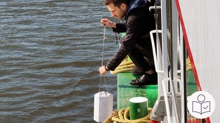 Ein Mann kniet am Rand von einem Boot. Der Mann hat ein Seil in der Hand. An dem Seil hängt eine Urne. Der Mann ist ein Bestatter bei einer Fluss·bestattung. Der Rechts das SWR Logo für Leichte Sprache.