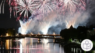 Ein Feuer·werk am Nacht·himmel. Das Feuer·werk ist über dem Bodensee. Es ist beim See·nachts·fest in der Stadt Konstanz. Menschen stehen auf zwei Brücken. Rechts das SWR Logo für Leichte Sprache.