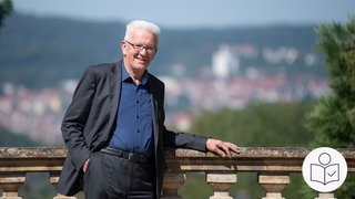 Das Bild zeigt Winfried Kretschmann. Er steht an einem Geländer. Im Hintergrund sieht man einen Garten und Häuser. Rechts das SWR Logo für Leichte Sprache.  