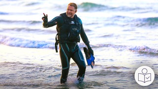 Der Engländer Karl Bushby läuft in einem Schwimm·anzug aus dem Meer. Mit einer Hand macht er das Victory-Zeichen für Erfolg. Rechts unten das SWR-Logo für Leichte Sprache.