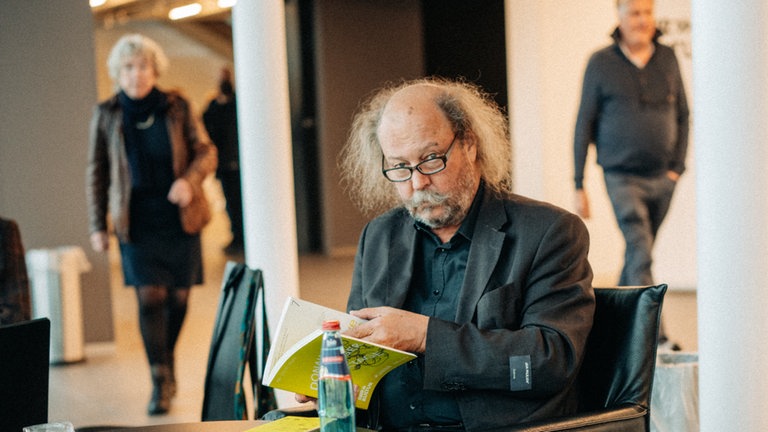 Sitzender Mann guckt von seinem Buch auf, direkt in die Kamera