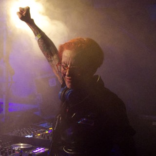 DJ Elly steht auf einer Bühne hinter dem DJ Mischpult. Sie streckt in Feierlaune eine Faust in die Luft. 