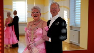 Christa und Karl-Heinz stehen Arm in Arm im Tanzsaal. Christa trägt ein langes, pinkes Tanzkleid. Karl-Heinz einen schwarzen Anzug. 