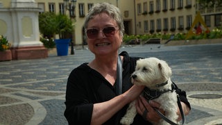 Eine ältere Dame mit grauen Haaren und Sonnenbrille mit einem kleinen weißen Hund auf dem Arm. 