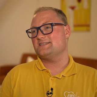 Junger Mann mit blonden Haaren, Brille und gelbem T-Shirt lächelt.