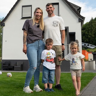 Junge Familie, Frau mit langen, hellen Haaren, grauen Shirt und Jeans. Mann mit kurzen, dunklen Haaren, weißem Shirt und zwei kleine Kinder.