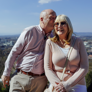 Jutta ist blond und trägt eine große Sonnenbrille. Klaus steht neben ihr und küsst sie auf die Schläfe. Hinter ihnen ist in der Ferne verschwommen Freiburg zu erkennen.