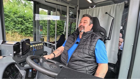 Busfahrer Pit sitzt in einem blauen T-Shirt und schwarzer Pufferweste in seinem Bus hinter dem Lenkrad. Sein Blick ist konzentriert nach vorne gerichtet. Im Hintergrund sieht man Bäume vorbeiziehen.