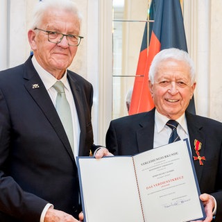 Für sein besonderes ehrenamtliches Engagement bekommt Helmut das Bundesverdienstkreuz verliehen. Auf dem Foto hält Helmut die Urkunde dazu in der Hand, neben ihm steht der baden-württembergische Ministerpräsident Winfried Kretschmann. Dieser übergibt Helmut die Urkunde. Hinter den beiden steht eine Deutschlandflagge und die Flagge von Baden-Württemberg. 
