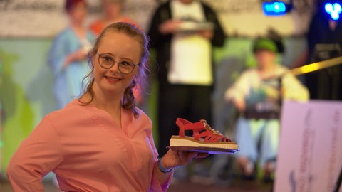 Eine junge Frau mit Down-Syndrom hält eine Sandale in die Kamera. Wiebke liebt es, auf der Bühne zu stehen und nimmt an einer inklusiven Modenschau teil.