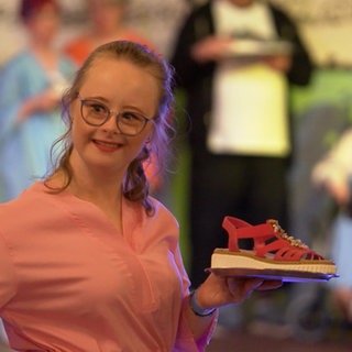 Eine junge Frau mit Down-Syndrom hält eine Sandale in die Kamera. Wiebke liebt es, auf der Bühne zu stehen und nimmt an einer inklusiven Modenschau teil.