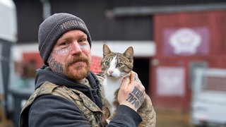 Mann mit Tattoos im Gesicht, dunkler Mütze und Katze auf dem Arm