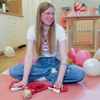 Julia sitzt auf dem Boden, im Schneidersitz, vor ihr liegt ein Plüsch-Modell eines Uterus und sie lächelt. Julia ist gerade dabei, einen Menstruationskurs zu geben, um sie herum liegen Luftballons und auch eine Teilnehmerin ist zu sehen.