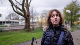 Manuela steht rechts im Vordergrund des Bilds. Sie trägt eine dunkle Winterjacke mit Kunstfellbesatz an der Kapuze. Man sieht das obere Ende ihres Blindenstocks. Sie steht auf dem Weg eines Parks.