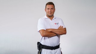 Notfallsanitäter Chris steht in seiner Rettungsdienst-Uniform vor einem grauen Hintergrund und lächelt in die Kamera.  