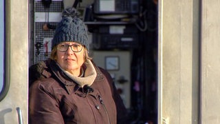 Fährfahrerin Martina steht auf der Fähre Neckarhausen. Sie trägt eine Mütze und eine Brille. 