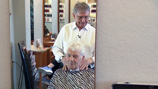 Günter ist 90 Jahre alt und Friseurmeister in Rente. Für seine Freunde und Bekannten kommt er aber gerne aus dem Ruhestand zurück. Günter steht hinter einem Kunden am Friseurstuhl. 