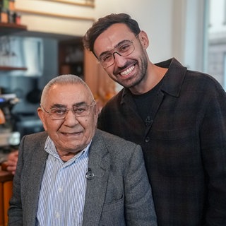 Barış und sein Opa Cemal stehen vor der Bar im Café Kalo. Barış hat ein Arm über die Schulter seines Opas gelegt und sie lachen in die Kamera.