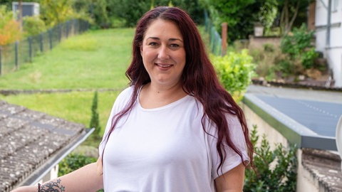 Junge Frau mit roten langen Haaren lächelt in die Kamera. Im Hintergrund sieht man einen grünen Garten.