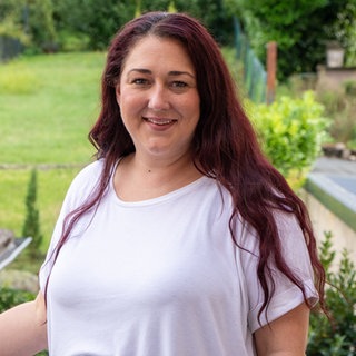 Junge Frau mit roten langen Haaren lächelt in die Kamera. Im Hintergrund sieht man einen grünen Garten.