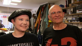 Eine junge Frau mit Ledermütze und ein älterer Mann mit Glatze. Beide tragen ein schwarzes T-Shirt mit Aufschrift.