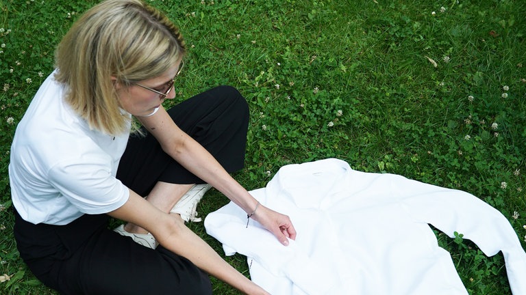Hannah sitzt auf einer großen Wiese. Vor ihr liegt eine weiße Bluse.