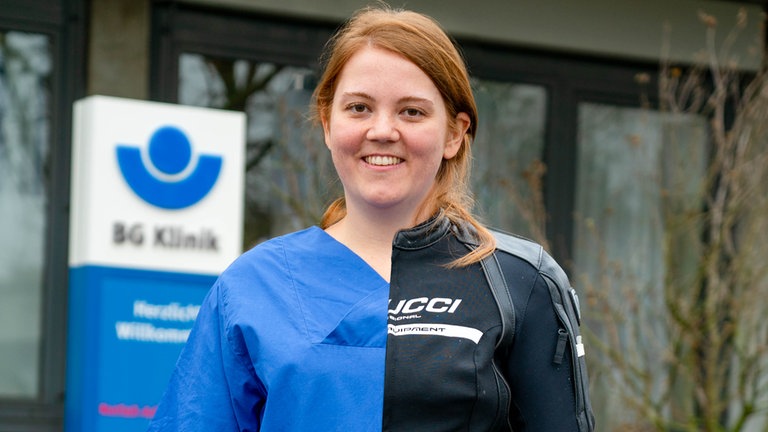 Junge Frau mit roten Haaren hat zur Hälfte einen blauen, medizinischen Kittel an, zur anderen Hälfte eine Motorrad Kombi.