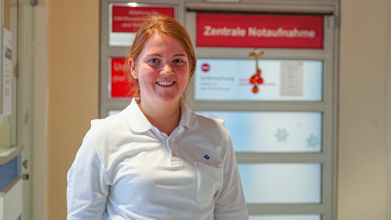 Junge Frau mit roten Haaren, weißem Polo-Shirt vor einer Tür mit der Aufschrift: Zentrale Notaufnahme.