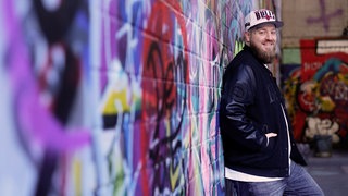 Junger Mann mit Bart und Mütze vor bunter Wand.