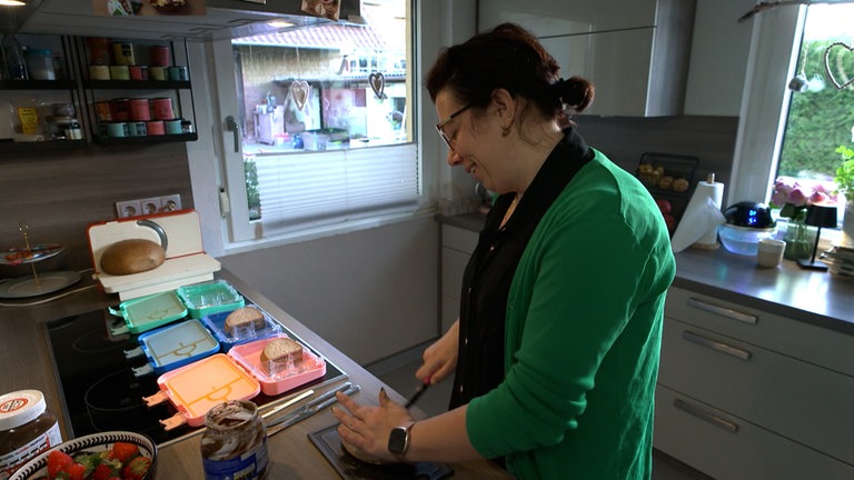 Eine Frau steht in der Küche vor drei Brotdosen und befüllt diese mit Obst und Broten.