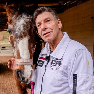Mann mit grauen Haaren und weißer Jacke steht mit einem Pferd vor einem Pferdestall.