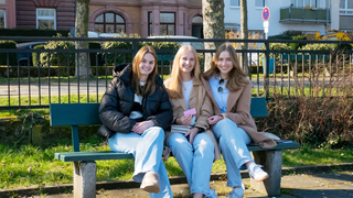 Drei junge Frauen sitzen auf einer PArkbank und lächeln in die Kamera