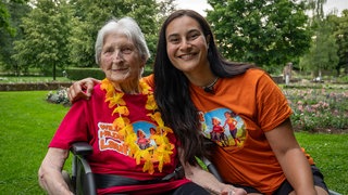 Seniorin im Rollstuhl sitzt neben einer jungen Frau in einem Park. Dei Seniorin hat kurze weiße Haare, trägt ein rotes T-Shirt mit dem Wilde Hilde Lauf Logo und eine Blumenkette. Die junge Frau hat lange, dunkle Haare, trägt ein orangenes T-Shirt mit Logo