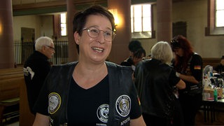 Frau mit kurzen Haaren und Brille, trägt eine Bikerkutte mit Abzeichen. Im Hintergrund weitere Frauen, die in einer Kirche zusammenstehen.