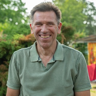Mann mit braunen Haaren trägt ein grünes Tshirt und lächelt in die Kamera. Im Hintergrund ist ein Wohnmobil zu sehen.