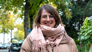 Junge Frau steht vor Bäumen und lächelt in die Kamera. 