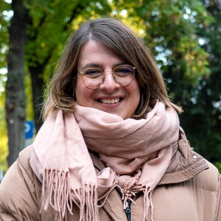 Junge Frau steht vor Bäumen und lächelt in die Kamera. 