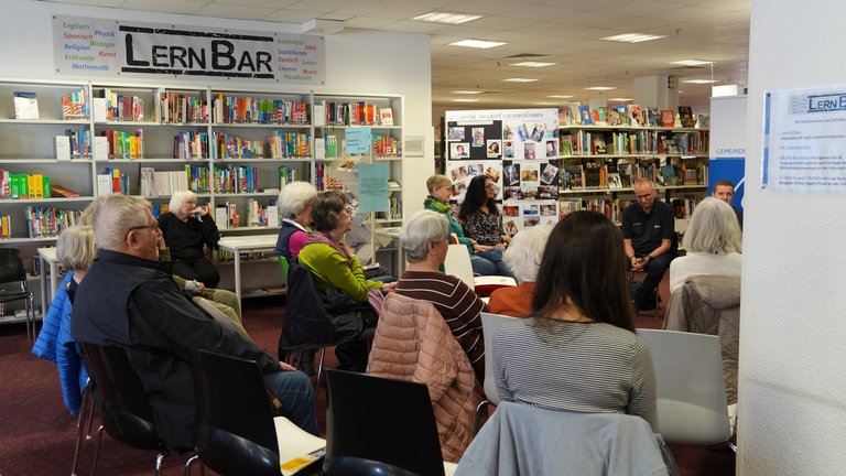 Viele ältere Menschen, die von hinten zu sehen sind. Sie sitzen in einer Bibliothek und schauen in eine Richtung. 