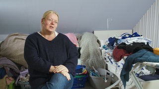 Eine blonde Frau mit Brille sitzt vor Wäschebergen.  