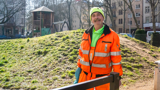 Mitarbeiter der Müllabfuhr. Trägt einen orangenen Anzug und eine grüne Mütze.