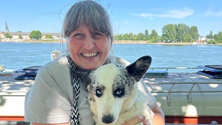 Ältere Frau mit grauen Haaren, hellem T-Shirt, Schal und Hund im Arm schaut in die Kamera. Hinter ihr ist ein Boot und dahinter sieht man den Rhein und die andere Uferseite.