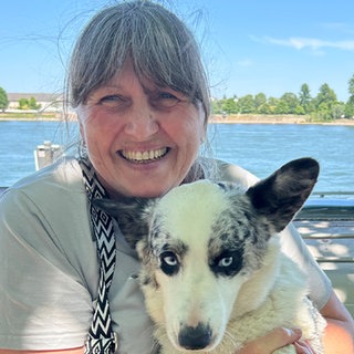 Ältere Frau mit grauen Haaren, hellem T-Shirt, Schal und Hund im Arm schaut in die Kamera. Hinter ihr ist ein Boot und dahinter sieht man den Rhein und die andere Uferseite.